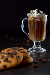 Close up of a glass cup of coffee with whipped cream and chocolate on it, chocolate chip cookies and roasted coffee beans on dark background. Concept of ready to eat food, tasty snack. Selective focus