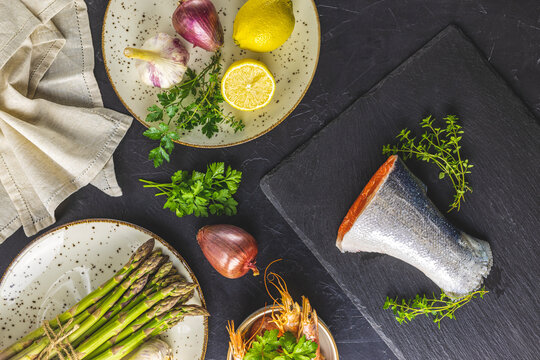 Trout fish surrounded parsley, lemon, shrimp, prawn, asparagus in ceramic plates. Black concrete table surface. Healthy seafood background..