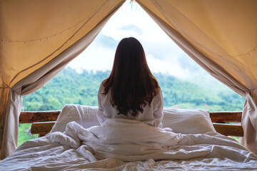 Rear view image of a woman sitting on the bed and looking at a beautiful nature view outside the tent