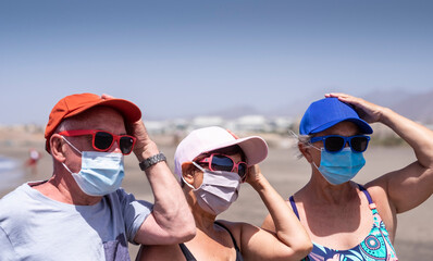 Group of three friends senior people enjoying together holidays at the beach under sun wearing face mask due to coronavirus - active retired elderly people and new normal concept