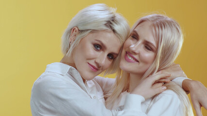 LGBT family portrait. Beautiful appealing blonde caucasian couple of girls in white shirts embrace and smile together against orange background. Copy space.