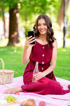 leisure and people concept - happy smiling woman with smartphone and fizzy drink in bottle sitting on picnic blanket at summer park
