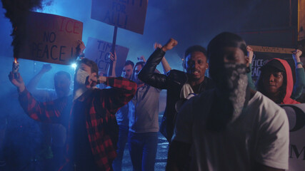 Striking crowd of multi-ethnic men and woman walking on street protest against police brutality with smoke gas grenades at night riot. Rebellion. Antiracism.