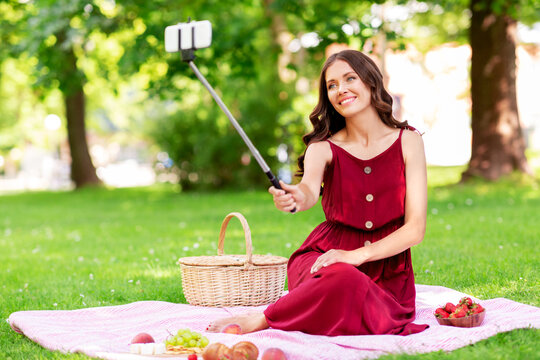 leisure and people concept - happy smiling woman with smartphone on selfie stick and picnic basket sitting on blanket and taking picture at summer park