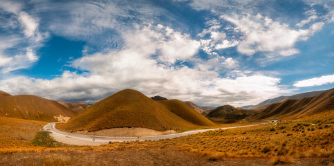 Naklejka na ściany i meble Panoramic view from a Desert Mountain Pass Summit with a two-lane highway curving through arid terrain. Golden light towards Lindis Pass in the summertime in Otago region, New Zealand, South Island.