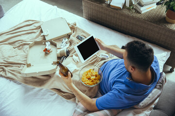 Eating crisps, drinks beer. Lazy man living in his bed surrounded with messy. No need to go out to be happy. Using gadgets, watching movie and series, looks emotional. Tablet with copyspace for ad.