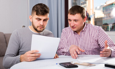 Two men in serious discussion analyzing documents together at home table, using phone