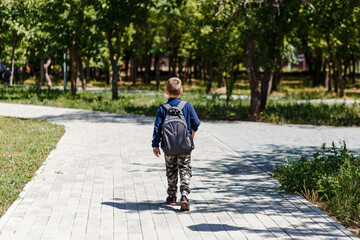 A schoolboy with a large backpack on his back walks along a path in the park.
