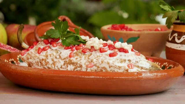 Chile en Nogada Mexican dish in traditional clay plate with pomegranate seeds and ingredients on wooden table, panning motion on food