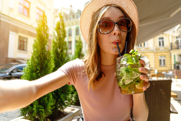 Young woman drinking mojito cocktail at cafe terrace at hot summer day and making selfie shot
