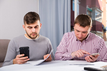 Two serious male friends reading attentively and discussing documents, using phone at home table