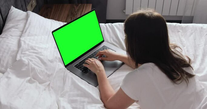 Young businesswoman lying on the bed working at day time on computer typing and thinking carefully looking at MacBook Pro 16 inch with keyed green screen on modern hotel room. Distance working.