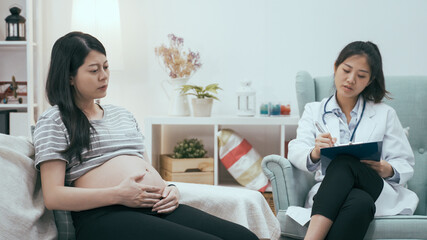 sad asian pregnant woman talking with female psychiatrist while sitting on sofa in clinic. depressed young motherhood complaining to mental medical staff. lady worker listening and make note.