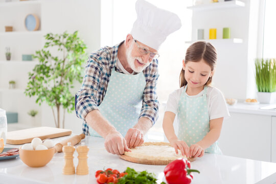 Portrait of nice cheerful cheery glad grey-haired granddad teaching grandchild learning cooking tasty yummy dish recipe national cuisine healthy nutrition in modern light white interior kitchen house
