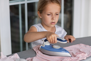Cute little girl with blonde hair is leaning on ironing clothes on board at home. Daughter helping...