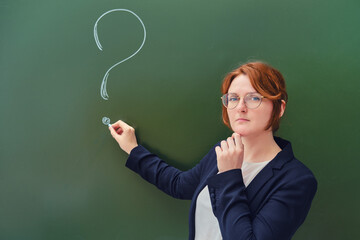 Teacher drew a question mark in chalk on a blackboard, copy space on green background. Woman teacher with a sad face at the school blackboard.