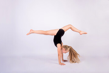 Fototapeta premium a little girl gymnast in a black swimsuit performs a handstand on a white isolated background with space for text