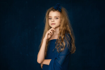Art Portrait of Beautiful young girl 10-12 years old with blonde curly long hair, holding her hands at her face on blue background