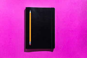 Orange pencil and black notepad from above on pink background. Office and school concept