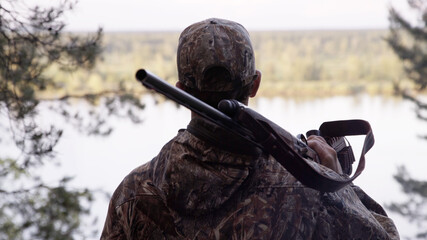 View from back of hunter in camouflage puts hunting rifle on his shoulder and looks at river from...