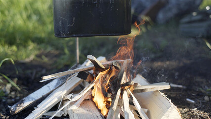 Black tourist pot hangs over burning flame of campfire in dark forest. Cooking dinner on wild hike. Cauldron of hot tea or food is heated over fire. Outdoor travel. Boil of hot meal in camp