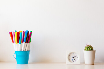 Many multi-colored felt tip pens in blue basket holder with little cactus and white small clock on the desk isolated white background, copy space