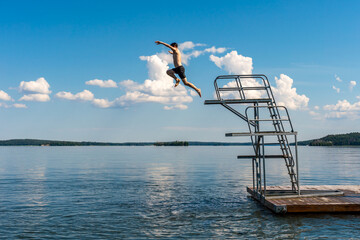 Side view of a teenage male jump diving from a diving tower with blue sky and horizon in the background.