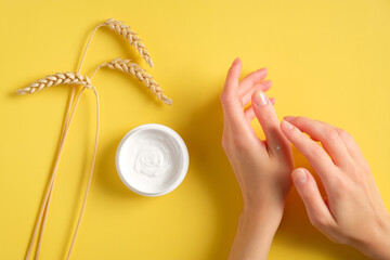Female hands applying wheat, barley and oat hand cream on yellow background. Natural skin care...