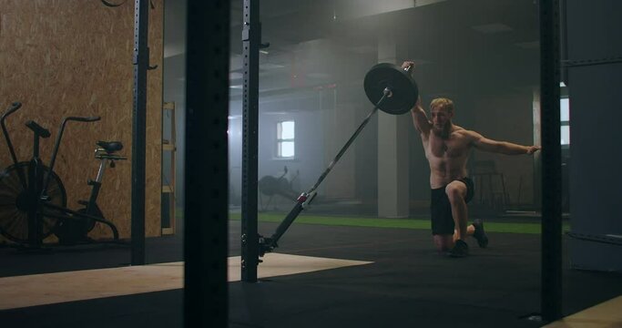 A man working out in the gym pushes a landmine bar with his hand while standing on one knee