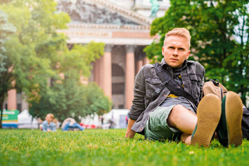 Naklejka na ściany i meble A young male tourist with a backpack visits the main sights of St. Petersburg, lying on the grass in front of St. Isaac's Cathedral