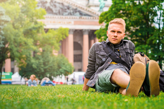A young male tourist with a backpack visits the main sights of St. Petersburg, lying on the grass in front of St. Isaac's Cathedral