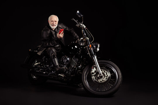 Full size photo of positive cool old man biker sit bike use smartphone search vacation adventure route navigation wear leather jacket modern clothes isolated over black color background