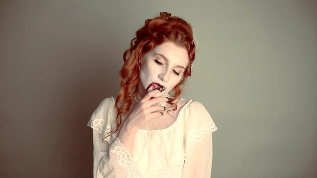 Young vampire woman with blood on face. Scary creepy character. Pale skin demon in white dress on gray background. Redhead model with red lips. Outfit for halloween. Vampire with open mouth and fangs