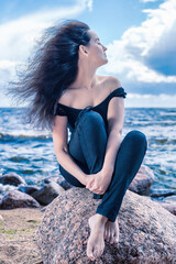 Calm brunette woman sitting on a stone by the sea with long hair flying in the wind