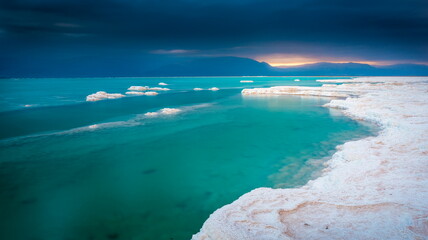 Beautiful Israeli landscape of the Dead Sea, Israel - the lowest place on Earth; turquoise-green water and white salt formations with dramatic clouds, as the sun rises over Jordan Mountains