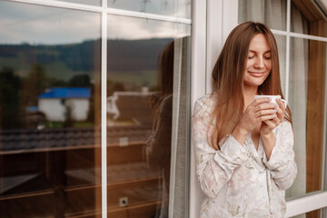 Young female standing after taking a shower in the morning on balcony of the hotel. holding a cup of coffee or tea in her hands. Looking outside nature forest and Mountain