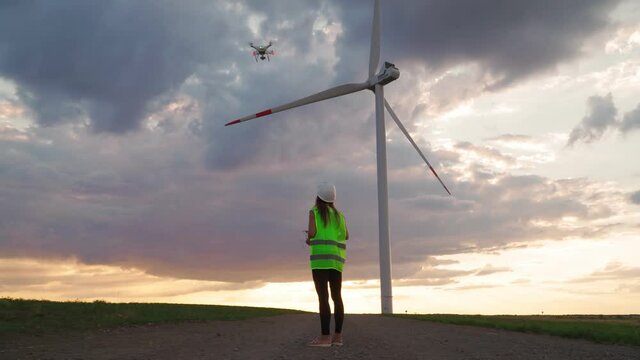 Woman Ecology engineer in uniform and helmet holding joystick controlling flying drone working at windmill on beautiful sunset background. Alternative to electrical energy