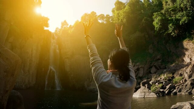 Woman standing alone raising up her hands before big waterfall at sunrise. traveling in Asian jungle. Healthy lifestyle and summer activity concepts.
