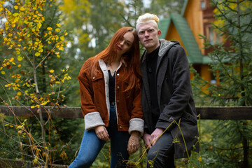Naklejka na ściany i meble A cute couple of young guys - a red-haired girl and an albino guy, stand huddled together at the fence against the background of the forest and a wooden house with a green roof