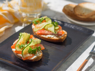 Open sandwiches with trout and cream cheese with cucumber and herbs on a dark plate - a tray on a gray table on a bright sunny day