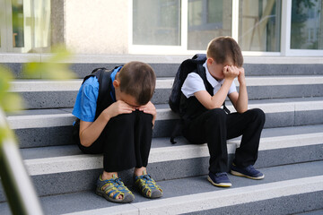 two sad boys, schoolchildren in black clothes are sitting on the steps of the porch of a building, covering their faces, the concept of fatigue, being late, punishment, back to school