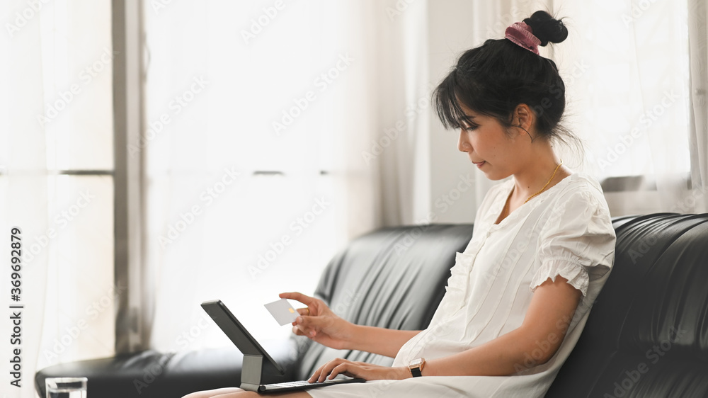Poster a beautiful woman is using a computer tablet and a credit card while sitting on the sofa.