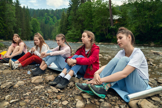 Female teens sitting on mats on river beach.