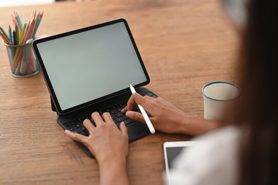 A cropped image of a woman is using a white blank screen tablet at the wooden table.