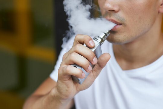 A young man standing on the street smokes a vape and blows smoke.