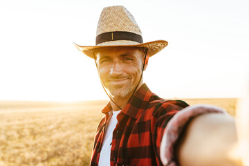 Fototapeta premium Image of pleased adult man in straw hat taking selfie photo while standing at cereal field