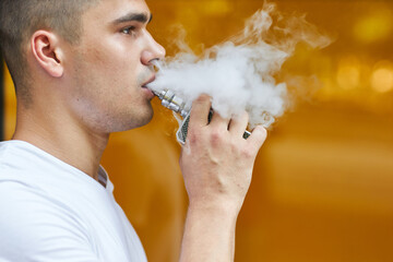 A young man standing on the street smokes a vape and blows smoke.
