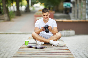 A young man sits with a laptop on a bench outdoors.
