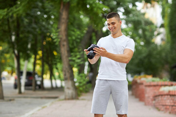 A young man stands outdoors with a camera and adjusts the camera