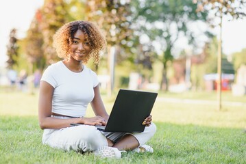 Young smiling black woman sitting outdoors on grass with laptop, typing, surfing internet. Technology, communication, education and remote working concept, copy space